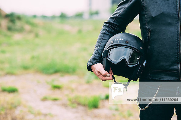Mid section of male motorcyclist standing on wasteland holding helmet