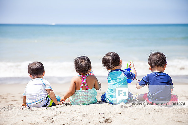 Rear view of baby girl and three baby boys sitting on beach Malibu California USA