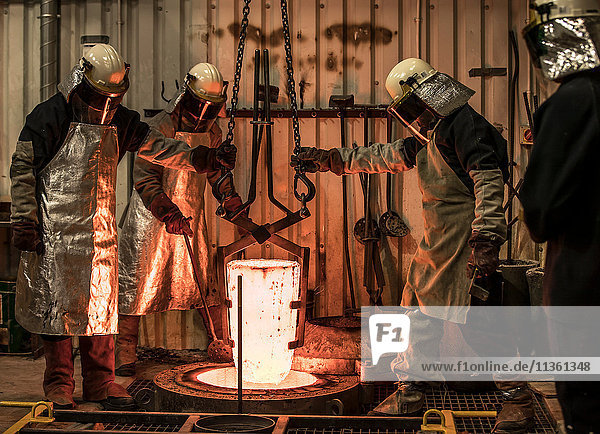 Male foundry workers winching white hot melting pot in bronze foundry