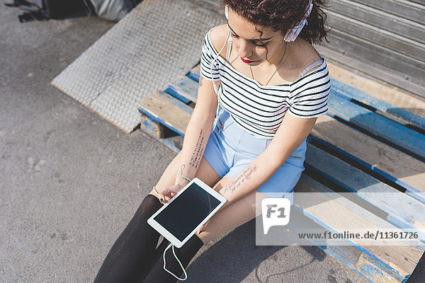 Woman sitting on pallet wearing headphones holding digital tablet