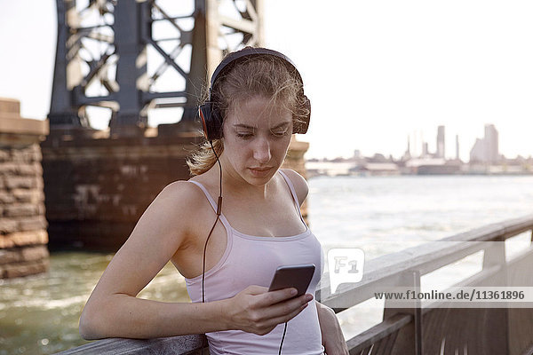Young woman beside river  wearing headphones  using smartphone  New York City  USA