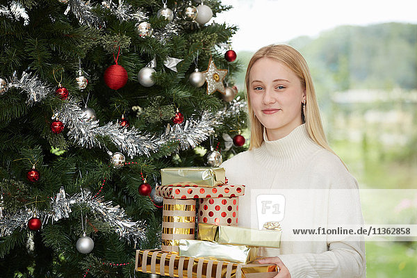 Porträt einer Teenagerin  die einen Stapel Weihnachtsgeschenke an einem Weihnachtsbaum hält