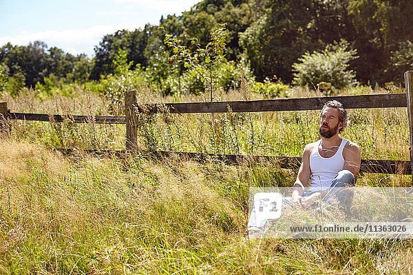 Mittelgroßer erwachsener Mann sitzt vor ländlichem Zaun und starrt