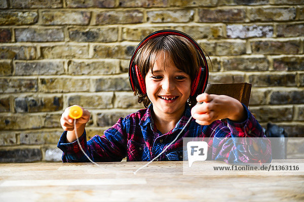 Glücklicher Junge sitzt am Tisch und spielt mit einem Spielzeug  während er Kopfhörer hört