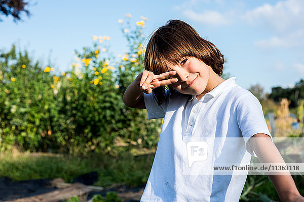 Porträt eines glücklichen Jungen  der im ländlichen Garten Friedenszeichen gibt