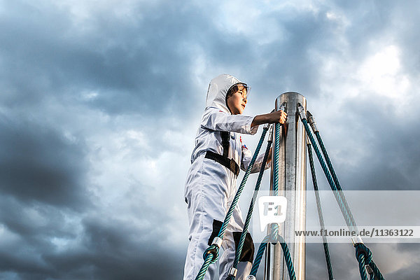 Junge im Astronautenkostüm blickt von der Spitze des Klettergerüsts gegen den dramatischen Himmel