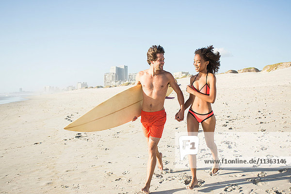 Junges Surferpaar läuft Hand in Hand am Strand  Kapstadt  Western Cape  Südafrika