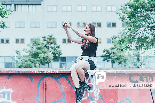 Junge Frau sitzt auf Graffiti-Wand und nimmt Smartphone-Selfie