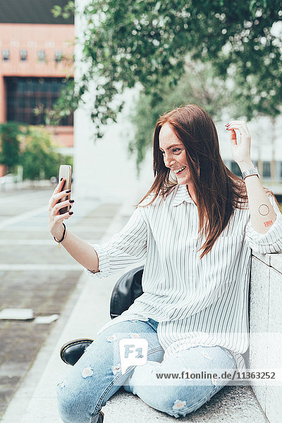 Junge Frau sitzt an der Wand und nimmt Smartphone Selfie