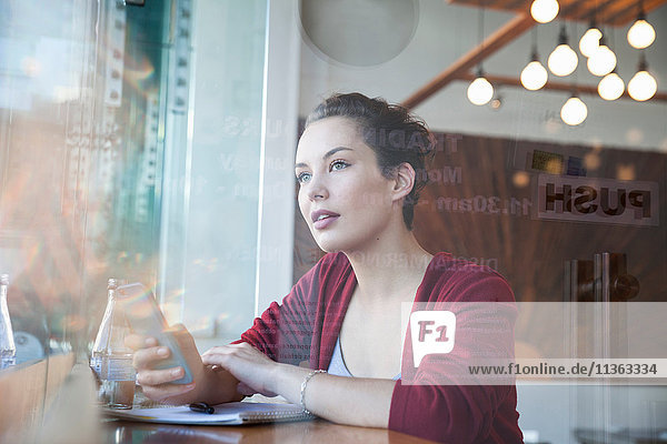 Junge Frau sitzt im Café  hält ein Smartphone in der Hand und schaut aus dem Fenster