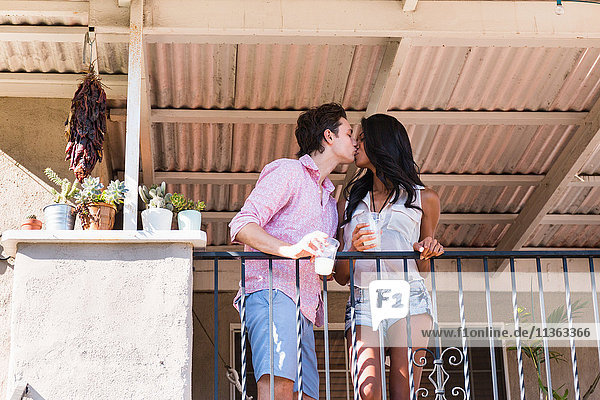 Junges Paar steht auf dem Balkon  hält Getränke in der Hand  küsst sich