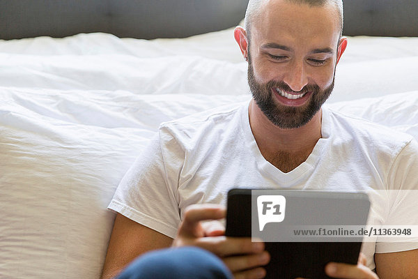 Mittelgroßer erwachsener Mann lehnt am Bett und stöbert auf digitalem Tablett