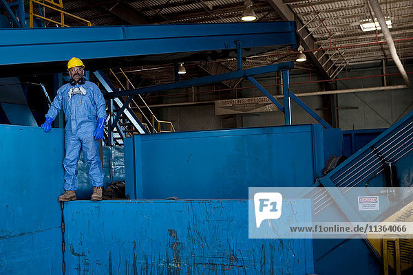 Porträt eines männlichen Arbeiters mit Schutzhelm  der auf blauen schweren Maschinen in einer Recycling-Anlage steht