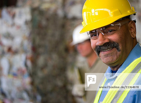 Porträt eines reifen männlichen Arbeiters mit Schutzhelm  vor gestapeltem Müll in der Recycling-Anlage