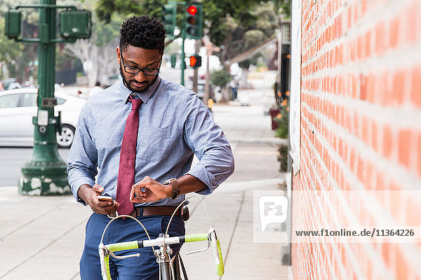 Junger Mann steht neben dem Fahrrad  benutzt ein Smartphone und schaut auf die Armbanduhr