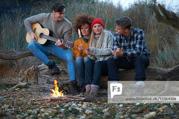 Vier junge erwachsene Freunde spielen Gitarre am Lagerfeuer am Strand in der Abenddämmerung