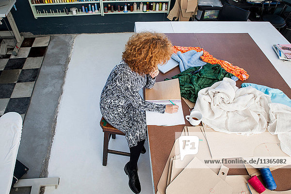 Hochwinkelansicht einer Zeichnung einer reifen Modedesignerin im Skizzenbuch am Werkstatttisch