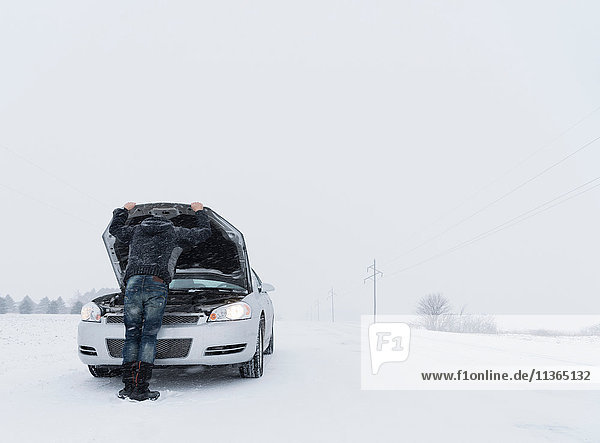 Rückansicht eines Mannes  der unter der Motorhaube eines liegen gebliebenen Autos auf eine schneebedeckte Landschaft schaut