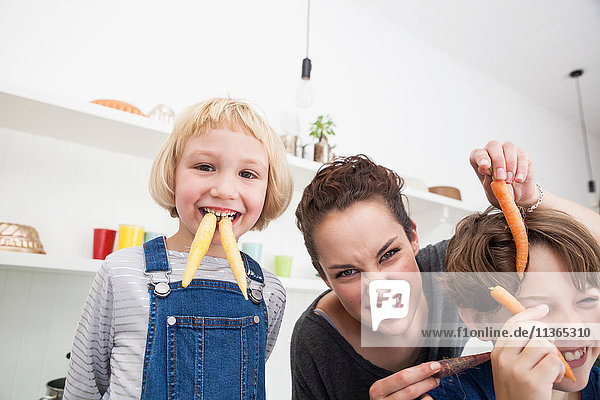 Young woman  boy and girl in kitchen  fooling around  using carrots as false teeth
