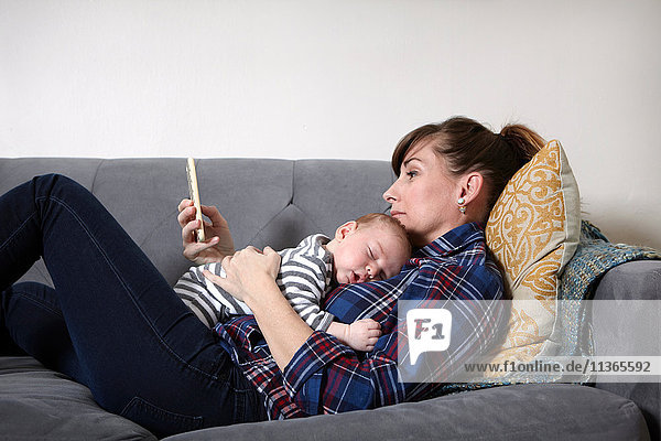 Mother with baby boy lying on sofa looking at smartphone
