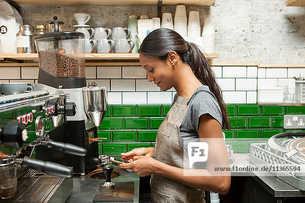 Weibliche Barista schaut auf Smartphone in Café-Küche