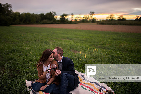 Romantic couple with red wine relaxing on picnic blanket in field at sunset