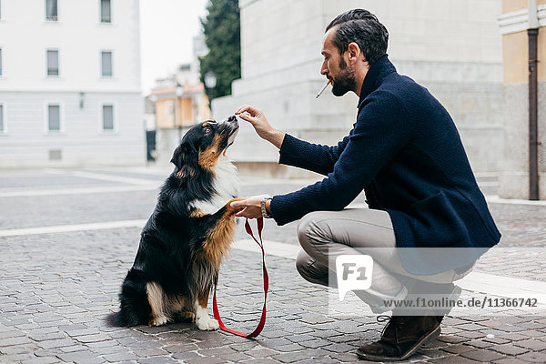 Mittelgroßer erwachsener Mann kauert mit seinem Hund auf dem gepflasterten Stadtplatz