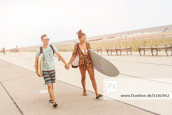 Junges Paar hält Hände und trägt Surfbretter am Rockaway Beach  New York State  USA
