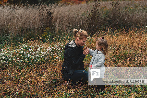 Mittelgroße erwachsene Frau  die an Wildblumen riecht  mit ihrer Kleinkind-Tochter in einem Feld mit langem Gras