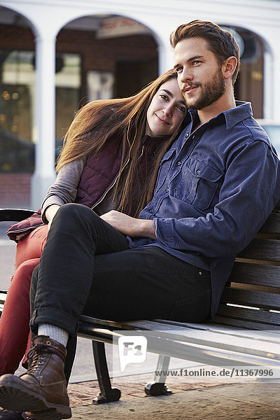 Ein junger Mann und eine junge Frau umarmen sich und sitzen auf einer Bank in einer städtischen Umgebung.