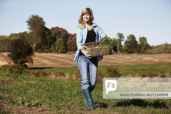 Eine junge Frau in Arbeitskleidung geht über ein Feld und hält einen Korb mit Feldfrüchten in der Hand.