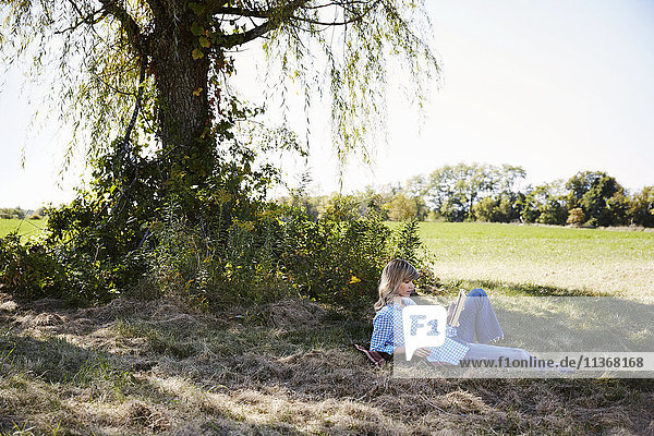 Eine Frau sitzt im Schatten eines Baumes in einer Bauernlandschaft und liest ein Buch.