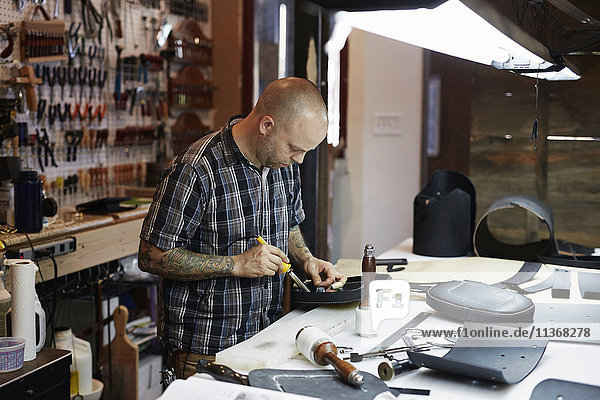 Ein Mann arbeitet an einer Bank unter hellem Licht in einer Lederwerkstatt. Eine große Werkzeugtafel mit Handwerkzeugen und Ausrüstung.