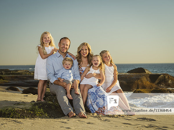 Familie mit Kindern (12-17 Monate  4-5  6-7  8-9) sitzend am Strand mit Meer im Hintergrund
