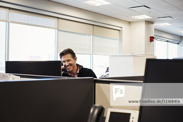 Ein Mann sitzt in einer Bürokabine mit einem Telefon am Ohr und lächelt.