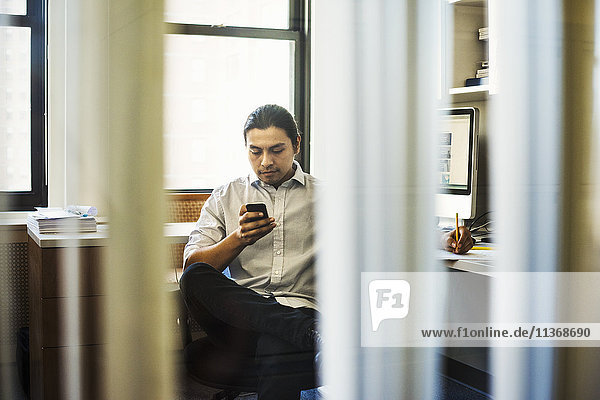 A man sitting on his own in an office checking his phone  seen through an open door.