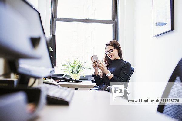Eine junge Frau sitzt an einem Schreibtisch in einem Büro und schaut auf ein Handy.
