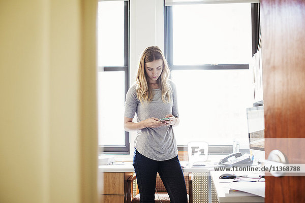 Eine junge Frau steht in einem Büro und schaut auf ihr Handy.