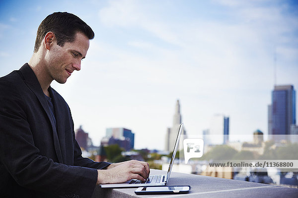 Ein junger Mann steht auf einem Dach und arbeitet an einem offenen Laptop mit einem Handy daneben.