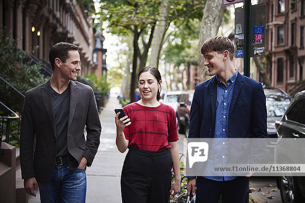 Two young men and a young woman walking down a city street looking at a cellphone.