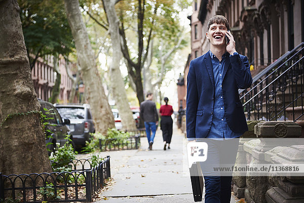 Ein junger Mann  der eine Straße in der Stadt entlanggeht und eine Aktentasche mit einem Mobiltelefon am Ohr hält