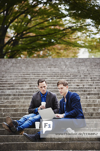 Two young men sitting on a flight of steps outdoors  looking at a laptop together.
