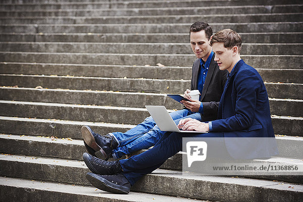 Zwei junge Männer sitzen auf einer Treppe und schauen gemeinsam auf einen Laptop.