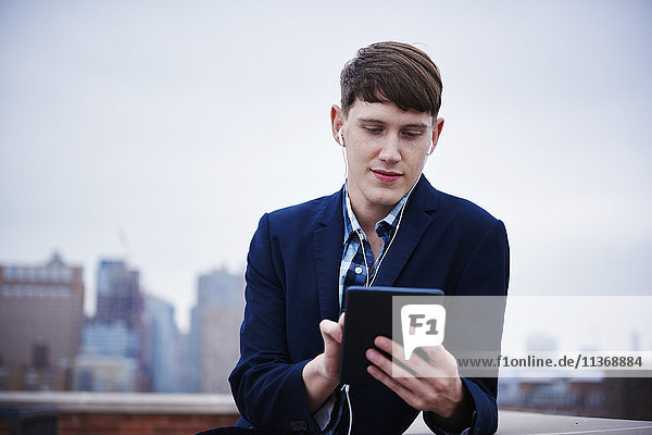 A young man standing on a rooftop looking down at a tablet.