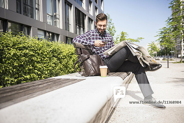 Junger Mann sitzt auf einer Bank und schreibt eine SMS auf seinem Smartphone