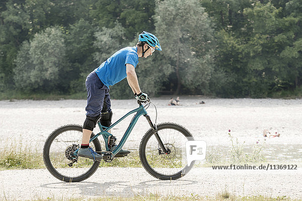 Mann fährt mit Mountainbike auf Pedalen stehend am Fluss entlang