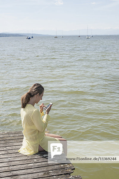 Frau benutzt Smartphone und sitzt auf Steg am See  Ammersee  Oberbayern  Deutschland