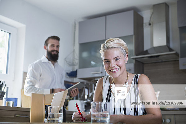 Junge Frau schreibt am Küchentisch und Mann im Hintergrund mit digitalem Tablet