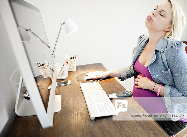 Schwangere Frau sitzt am Schreibtisch und fühlt sich krank