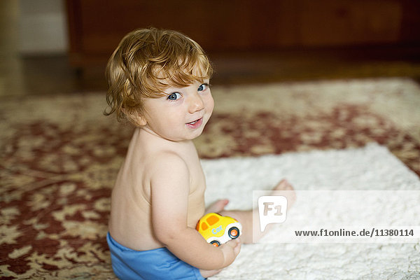 Kaukasischer kleiner Junge sitzt auf einem Teppich und hält ein Spielzeugauto